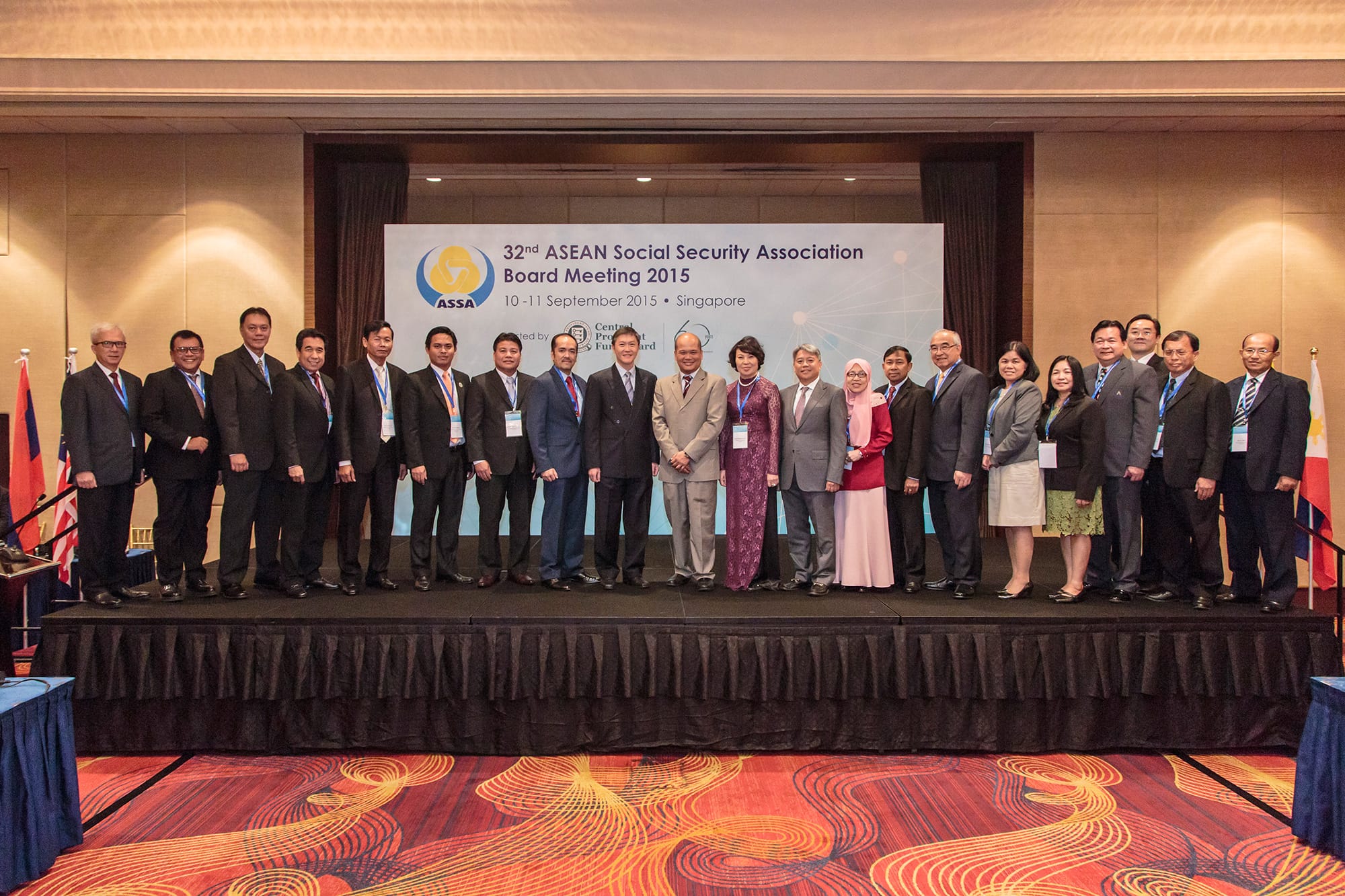 A group of delegates pose for a formal photograph at the 32nd ASEAN Social Security Association (ASSA) Board Meeting held in Singapore on 10–11 September 2015, as indicated by the backdrop behind them. The meeting was hosted by the Central Provident Fund (CPF) Board, which was also celebrating its 60th anniversary, as reflected in the logo on the backdrop. The participants, dressed in formal attire, are standing on a raised stage with ASEAN member country flags partially visible on either side.